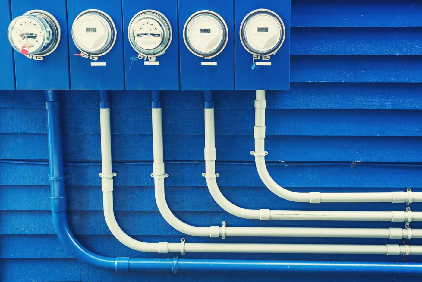 A row of electric meters mounted on a vibrant blue wall with neatly routed conduits.