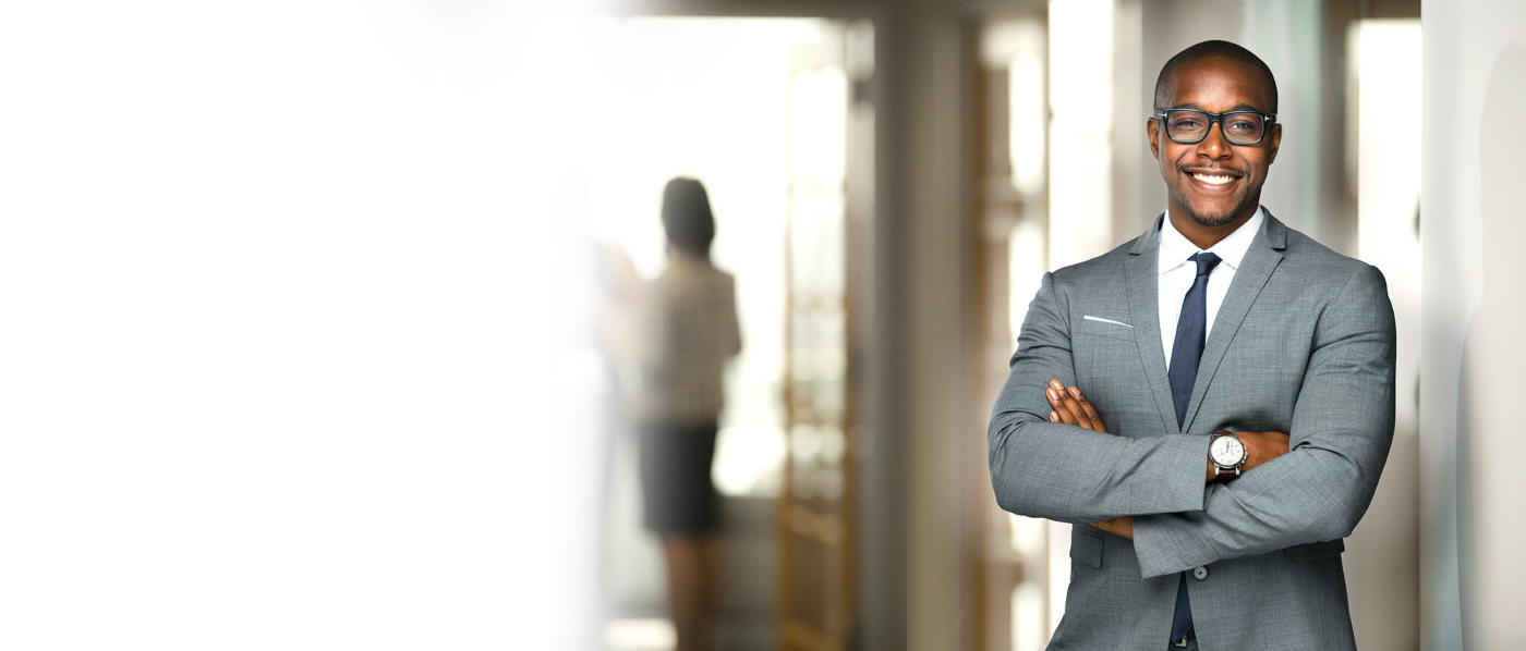A smiling business professional stands with arms crossed in a bright office corridor.