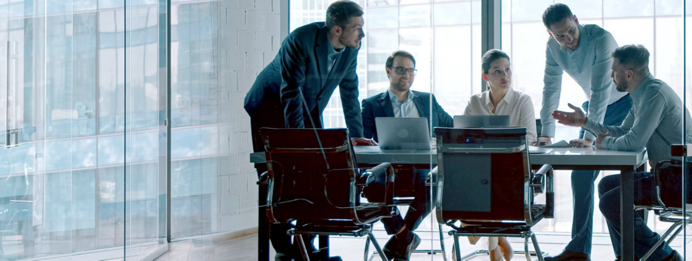 Colleagues gather around a table with laptops during a meeting in a glass-walled office.