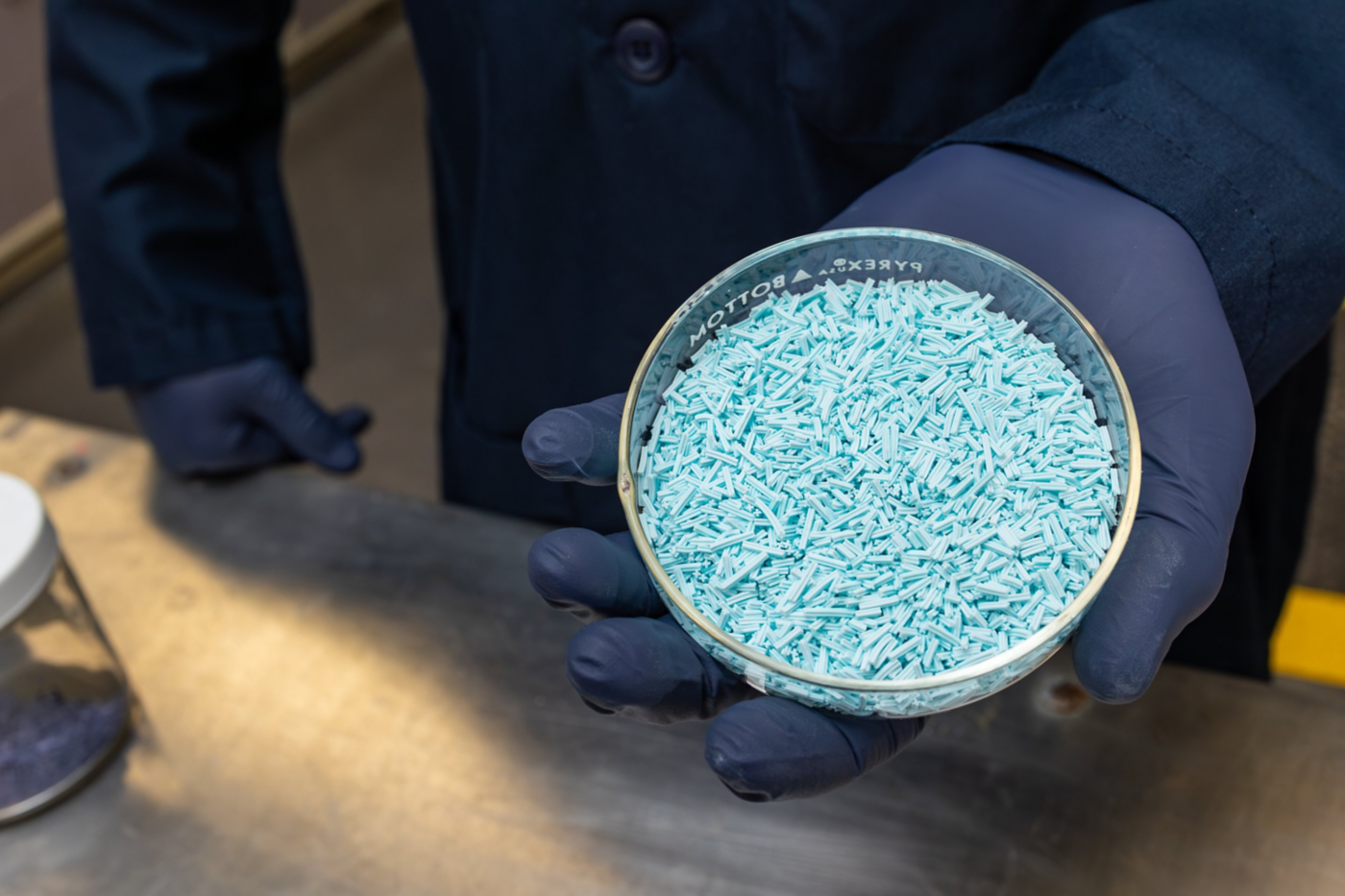 A technician in a lab coat and gloves holds a petri dish filled with light blue cylindrical chemical catalyst pellets for industrial quality inspection.
