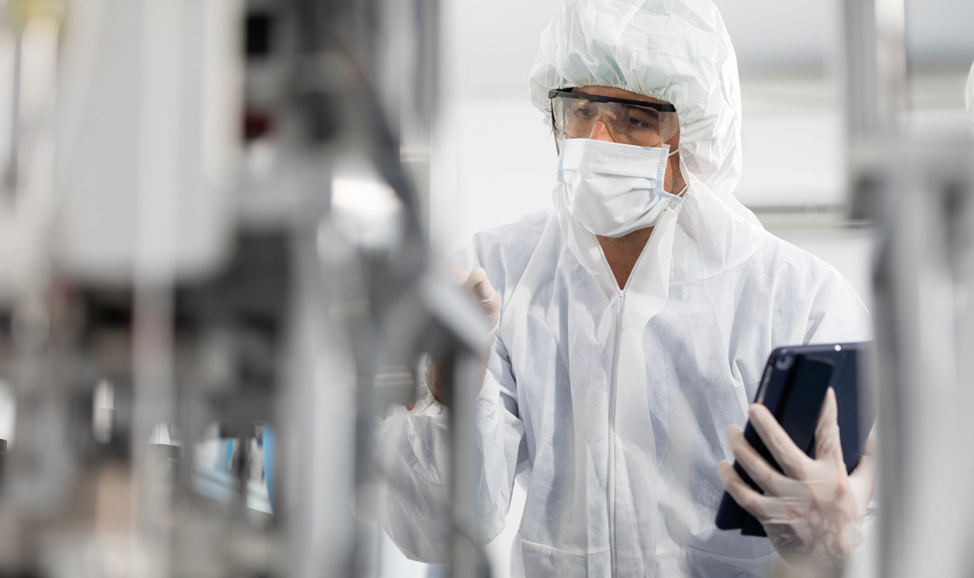 A masked worker in white cleanroom clothing holds a tablet inside a pharmaceutical production facility.
