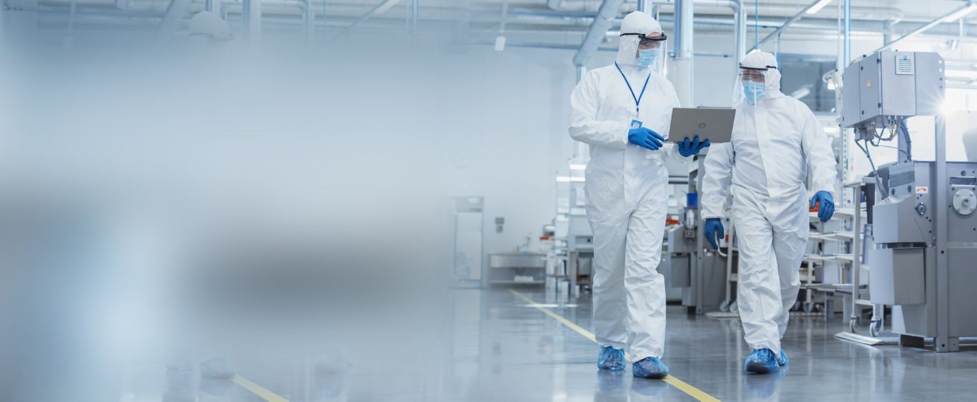 Two workers in full protective suits walk through a cleanroom while one carries a tablet.