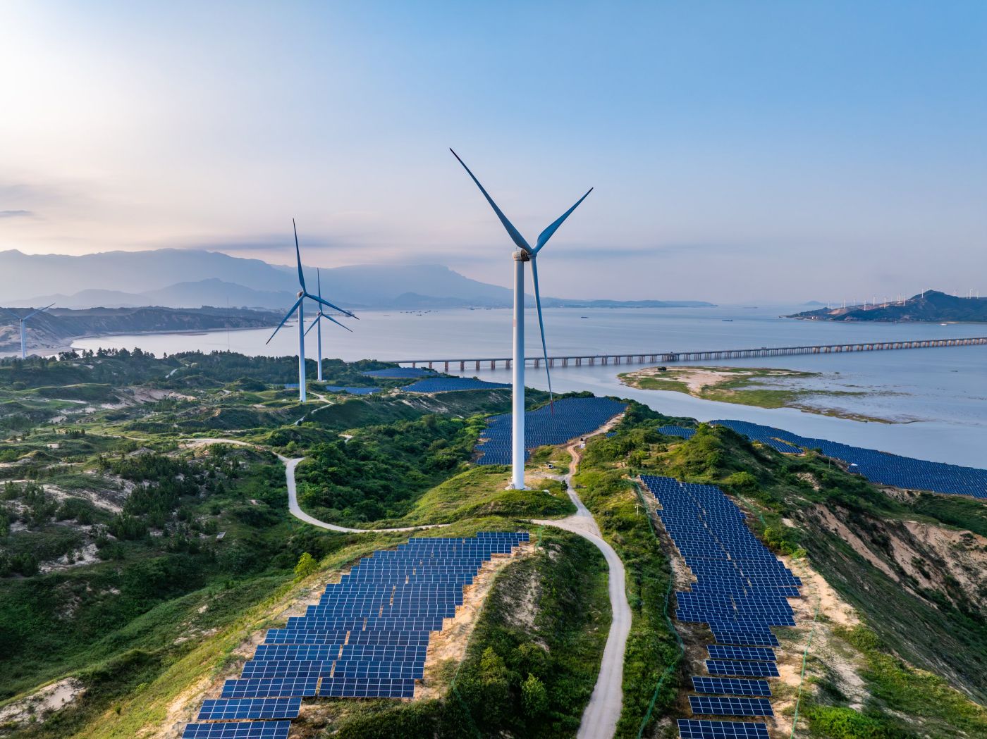 Aerial view of a hillside solar farm integrated with wind turbines overlooking a calm coastal bay and bridge.