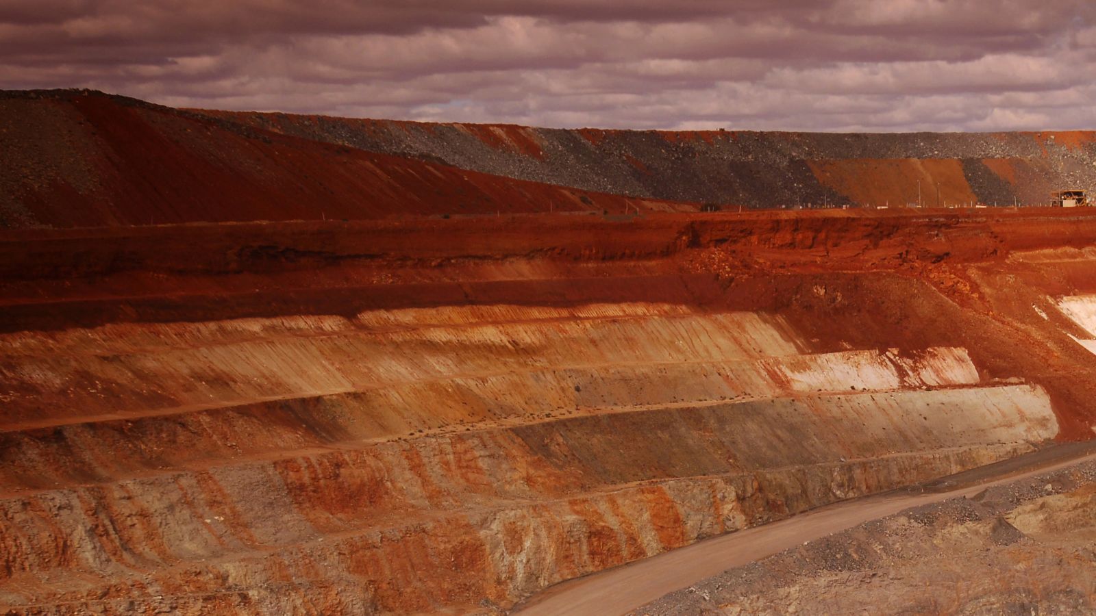 Wide view of an open pit mine showing layered earth, stepped slopes, access road, and overcast sky.
