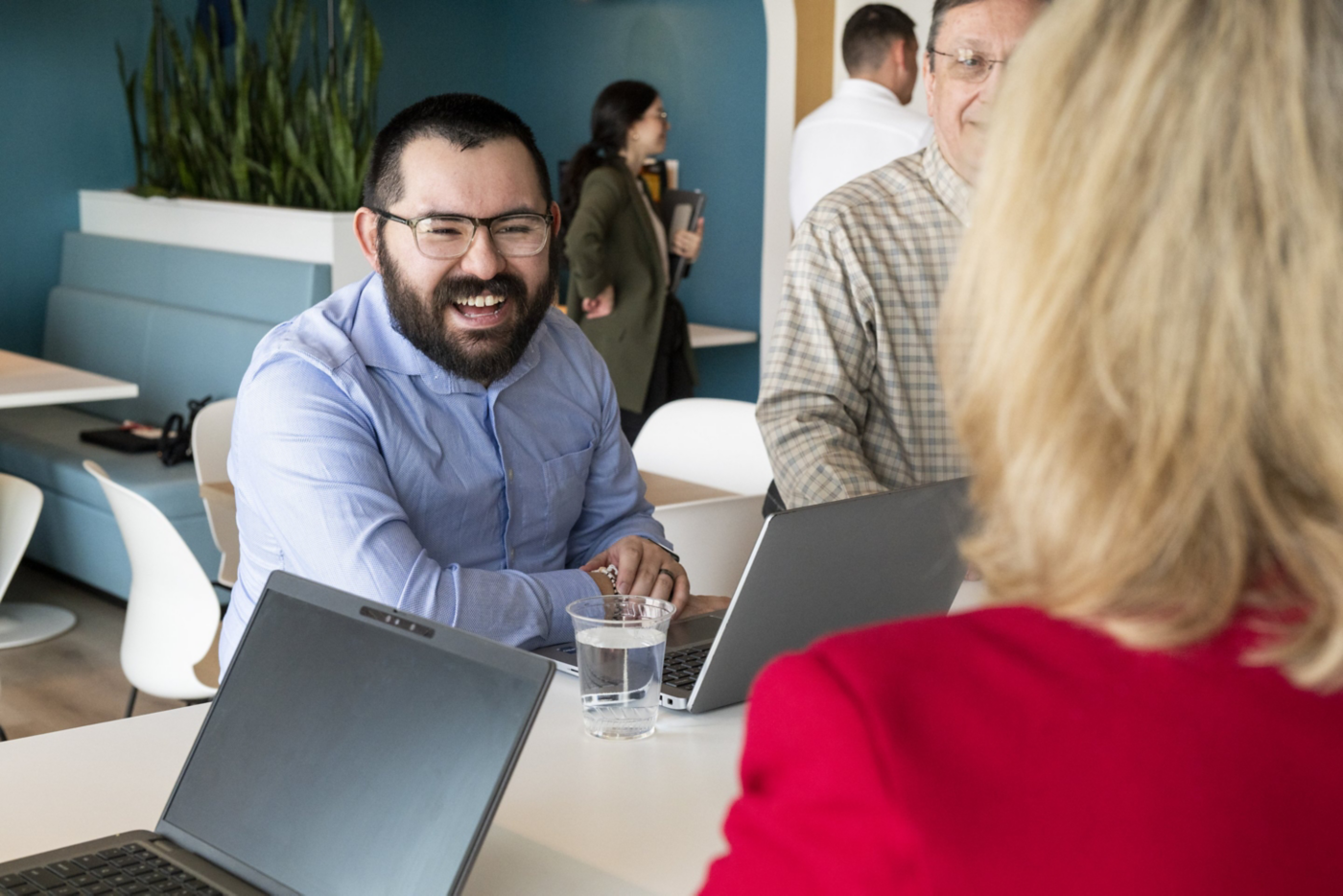 A smiling man in a blue shirt engages with coworkers during a collaborative meeting in a bright, modern corporate office environment.