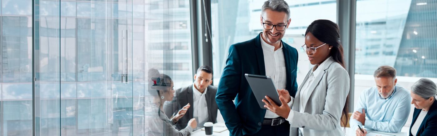 Two business colleagues review a tablet while other professionals meet in a glass-walled office.