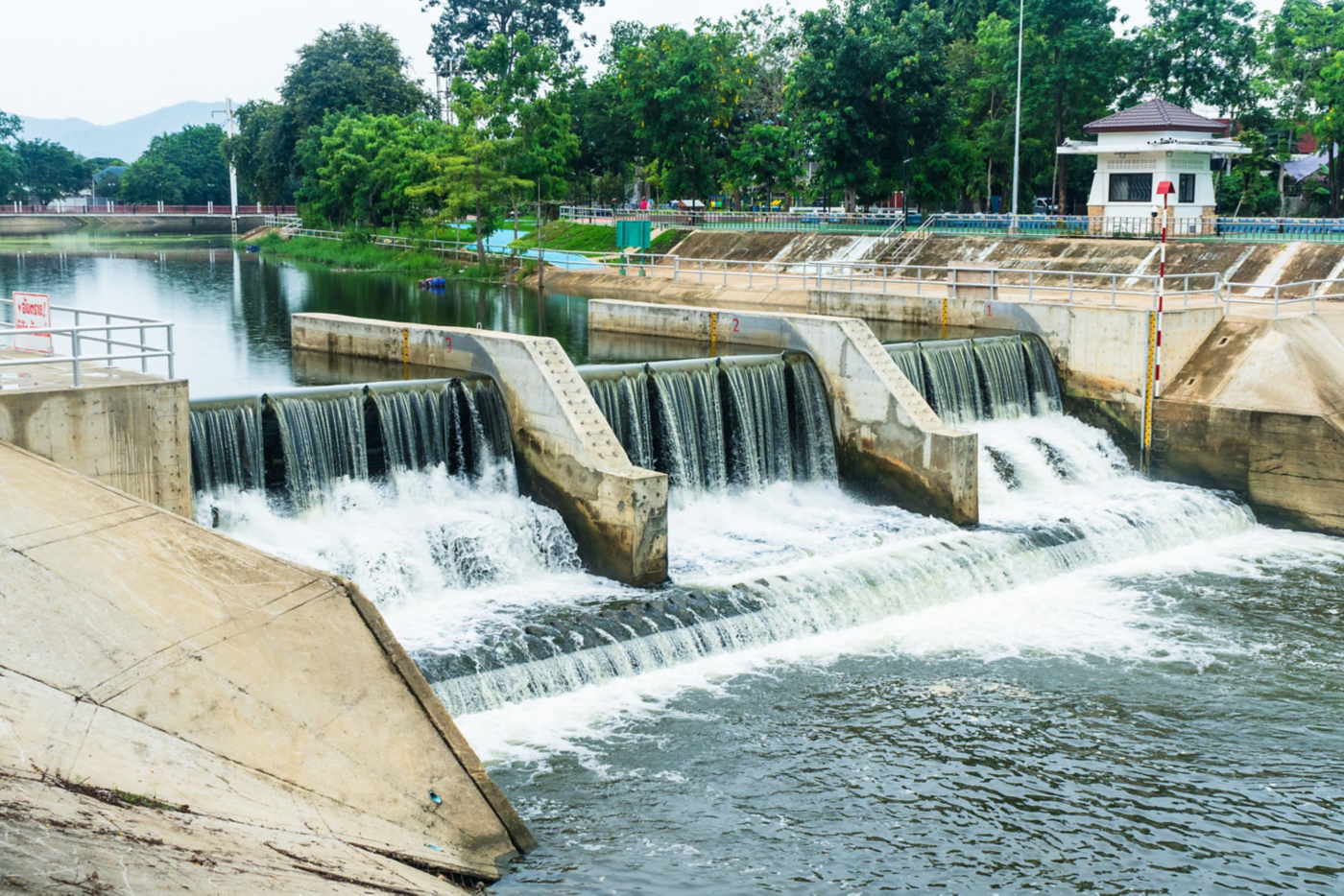 A concrete water diversion weir features multiple cascading sections creating a small waterfall within a landscaped public park setting.