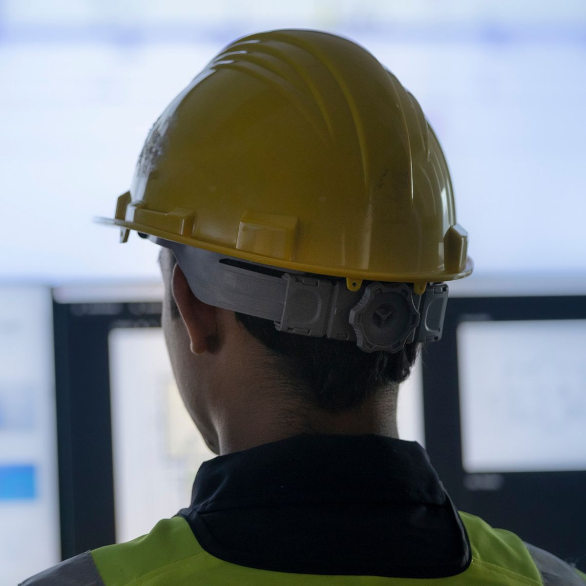 A worker wearing a yellow safety helmet and high-visibility vest stands facing multiple large computer monitors displaying blurred dashboards and data interfaces, suggesting industrial system monitoring or control in a technical environment.