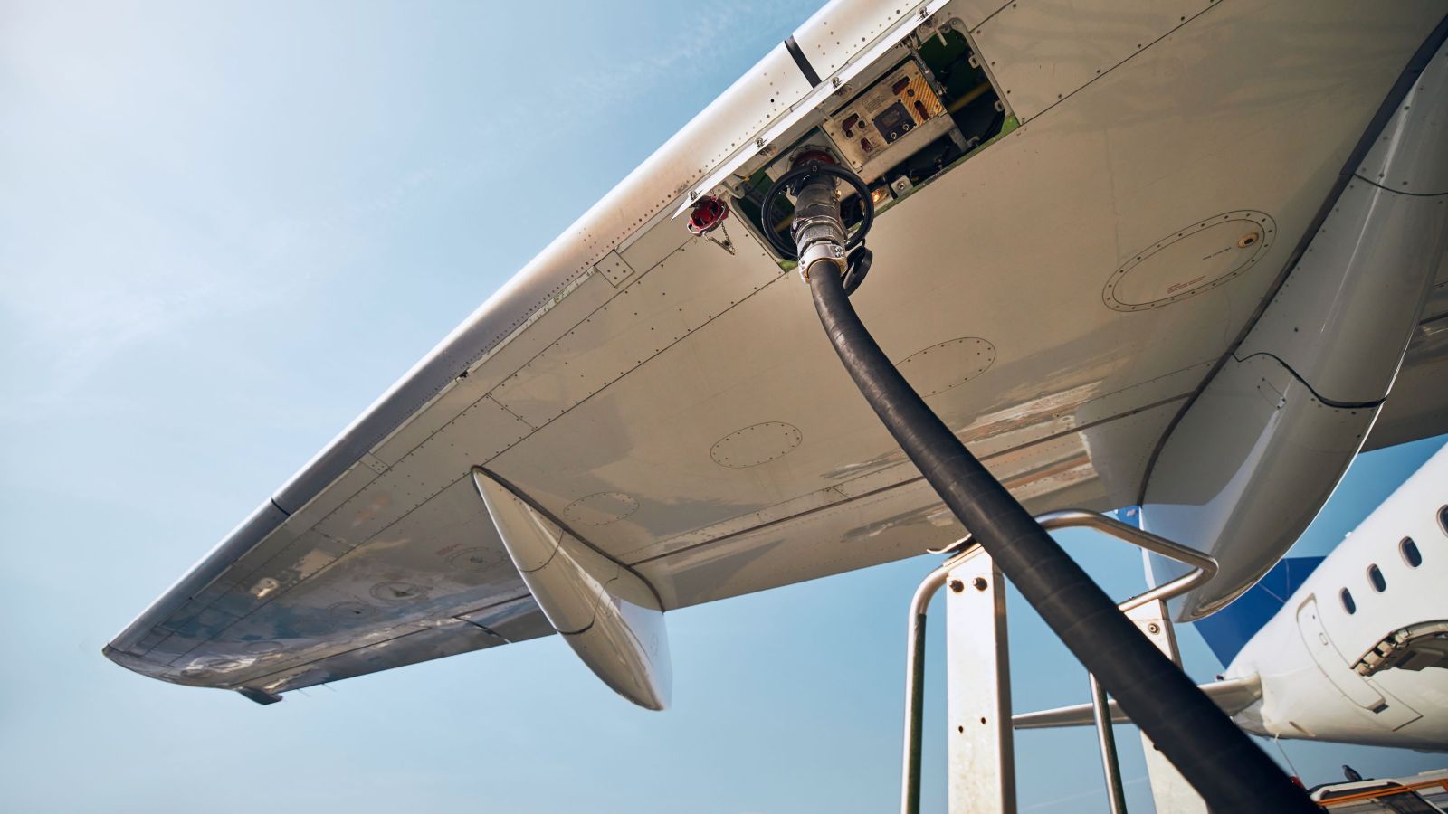 An airplane wing with a fuel hose attached for refueling, showcasing the aircraft against a bright blue sky.