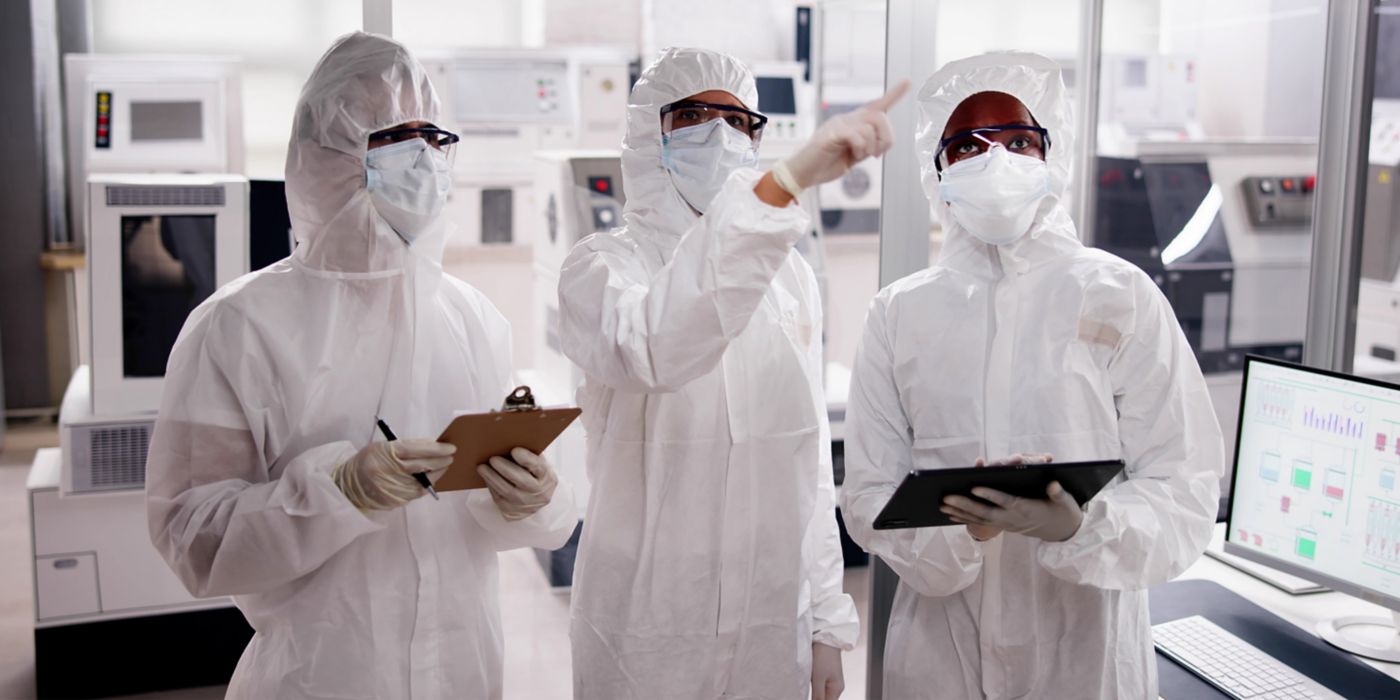 A group of three researchers wearing white protective suits and gloves collaborate in a laboratory setting. One researcher is holding a clipboard while another points toward a screen displaying data, indicating a technical discussion. The background features laboratory equipment, illustrating a high-tech scientific environment.