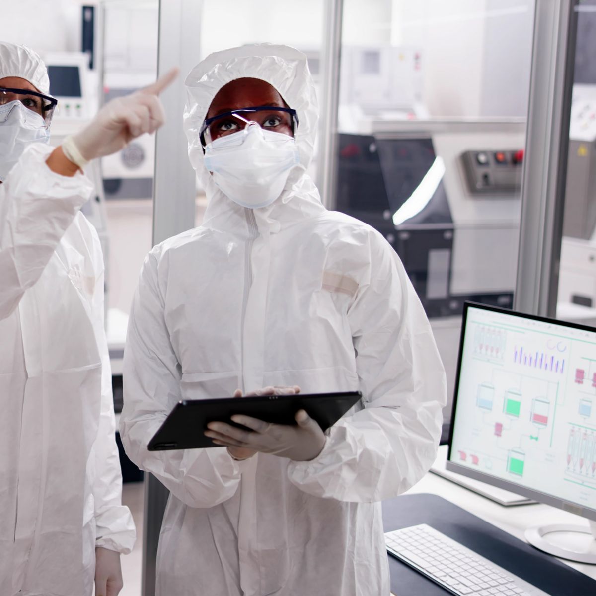 A group of three researchers wearing white protective suits and gloves collaborate in a laboratory setting. One researcher is holding a clipboard while another points toward a screen displaying data, indicating a technical discussion. The background features laboratory equipment, illustrating a high-tech scientific environment.
