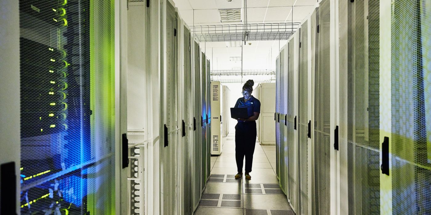 A professional works in a data center, surrounded by server racks while using a laptop. Soft blue and green lighting creates a high-tech atmosphere.