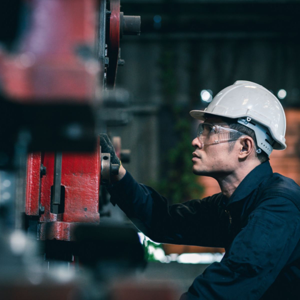 A worker wearing a hard hat and safety glasses adjusts a red industrial machine component in a factory setting.