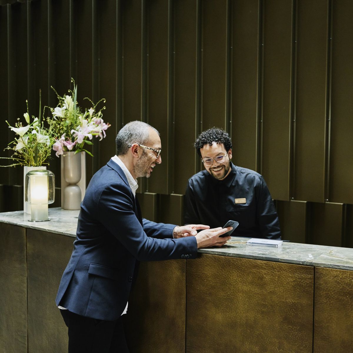A professional concierge and businessman share a friendly interaction at a modern luxury hotel reception desk.