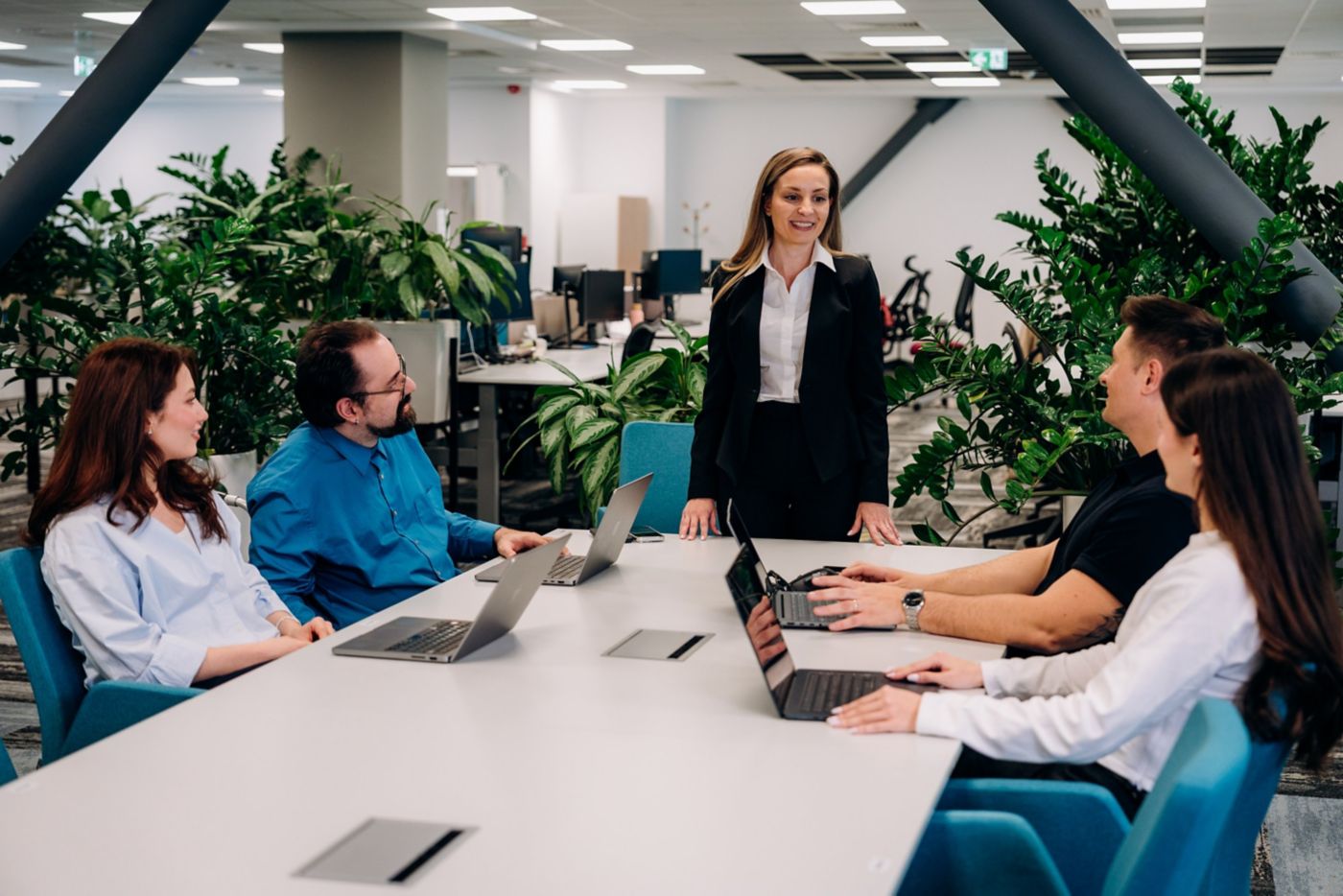 A confident female leader stands at the head of a modern office conference table, engaging her professional team during a collaborative business strategy session.