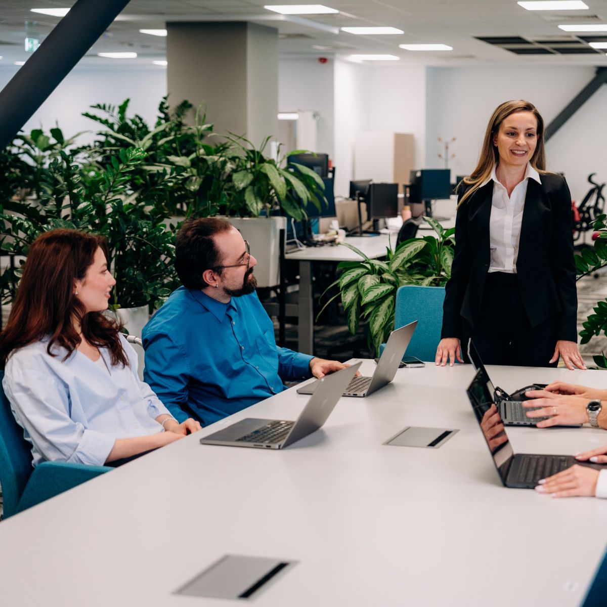 A confident female leader stands at the head of a modern office conference table, engaging her professional team during a collaborative business strategy session.