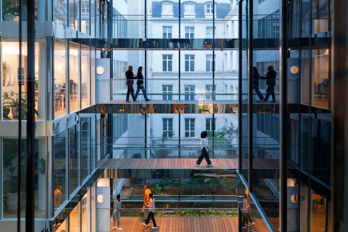 Employees navigate multi-level glass walkways in a modern office atrium featuring internal greenery and reflective architecture.