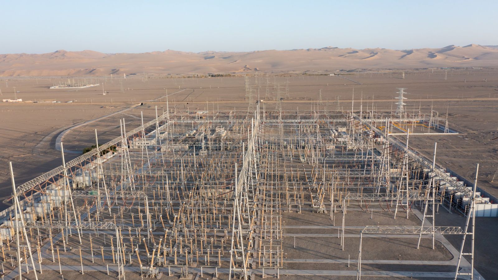 An aerial view captures a large-scale industrial electrical substation situated within a vast, arid desert landscape under a clear sky.