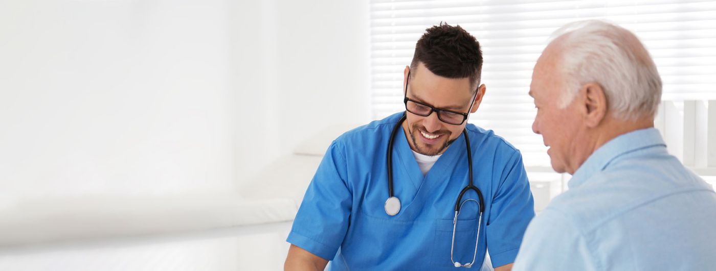A doctor in blue scrubs smiles while speaking with an older male patient in a bright clinical room.
