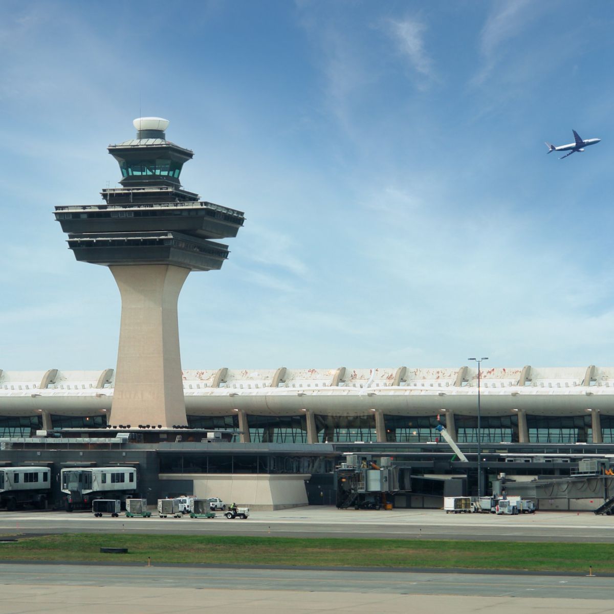 An iconic mid-century modern air traffic control tower stands prominently at Dulles International Airport as an airplane ascends into the blue sky.