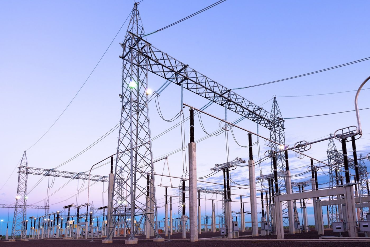 Steel transmission towers and high-voltage electrical equipment stand against a clear dusk sky at a utility power substation.