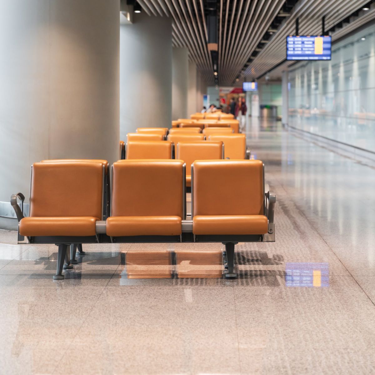 Rows of empty orange seats in a modern, minimalist airport terminal with reflective floors and architectural lighting.