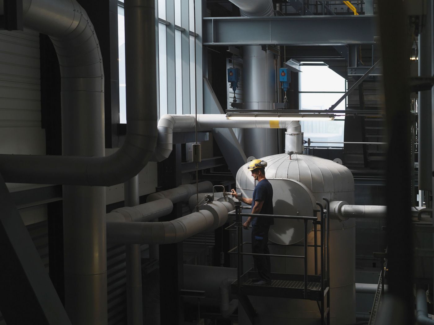 A worker stands on a platform, inspecting equipment in an industrial space filled with pipes and machinery.