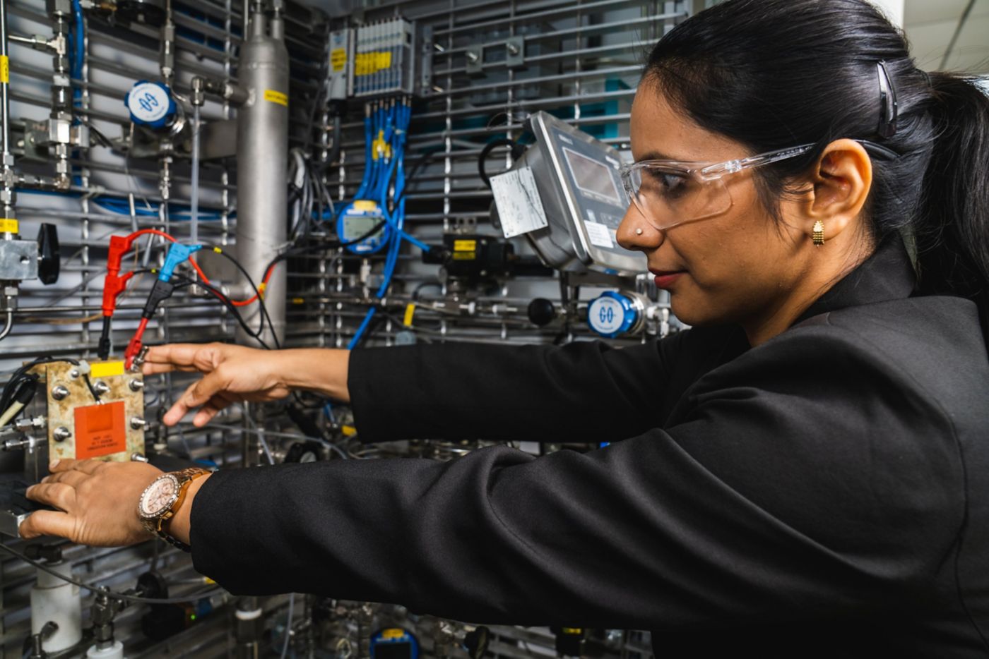 An engineer in safety glasses meticulously inspects complex laboratory hardware with wires and gauges.