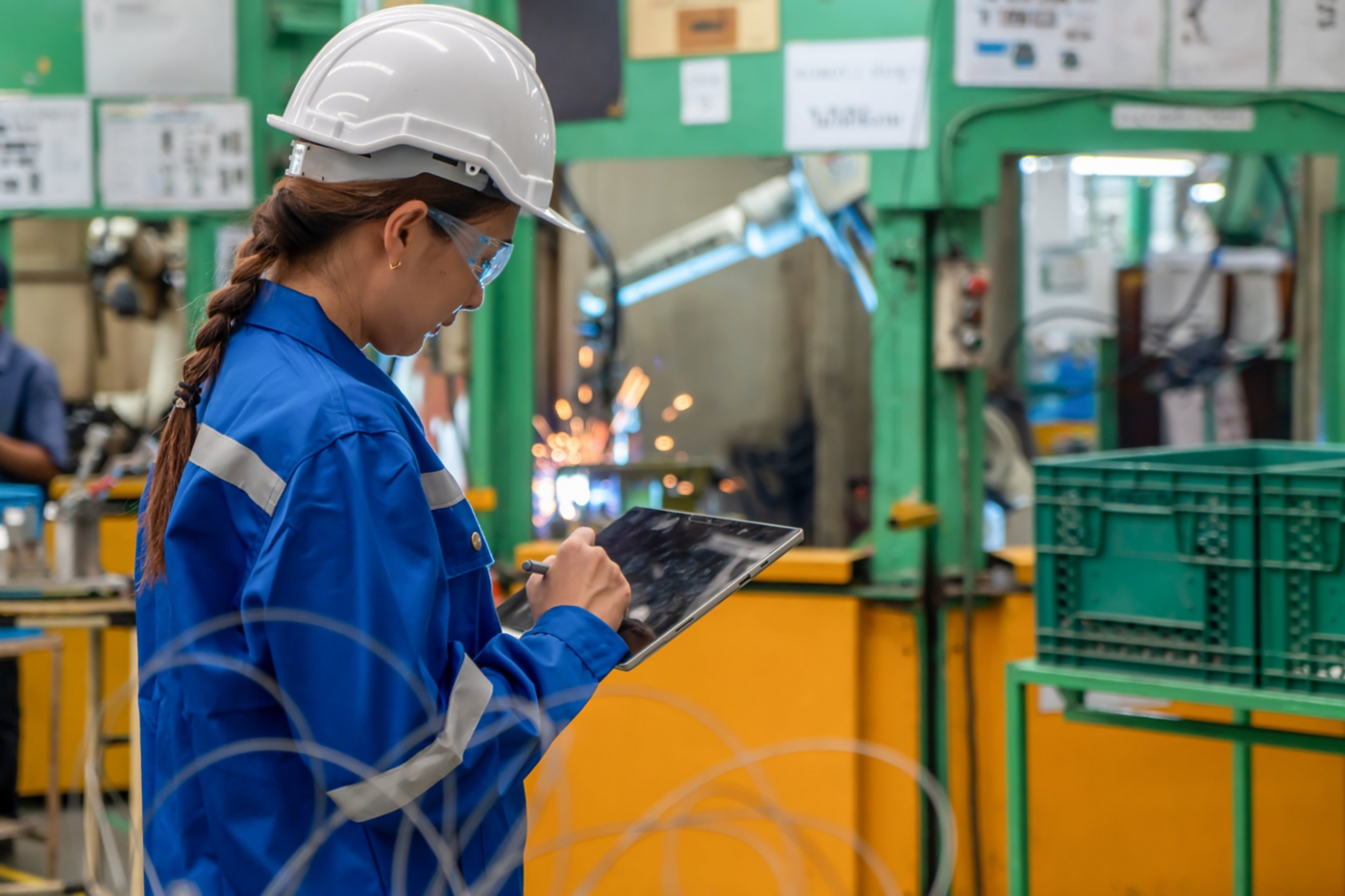Female engineer in safety gear uses a tablet to monitor manufacturing processes while industrial sparks fly in the blurred factory background.