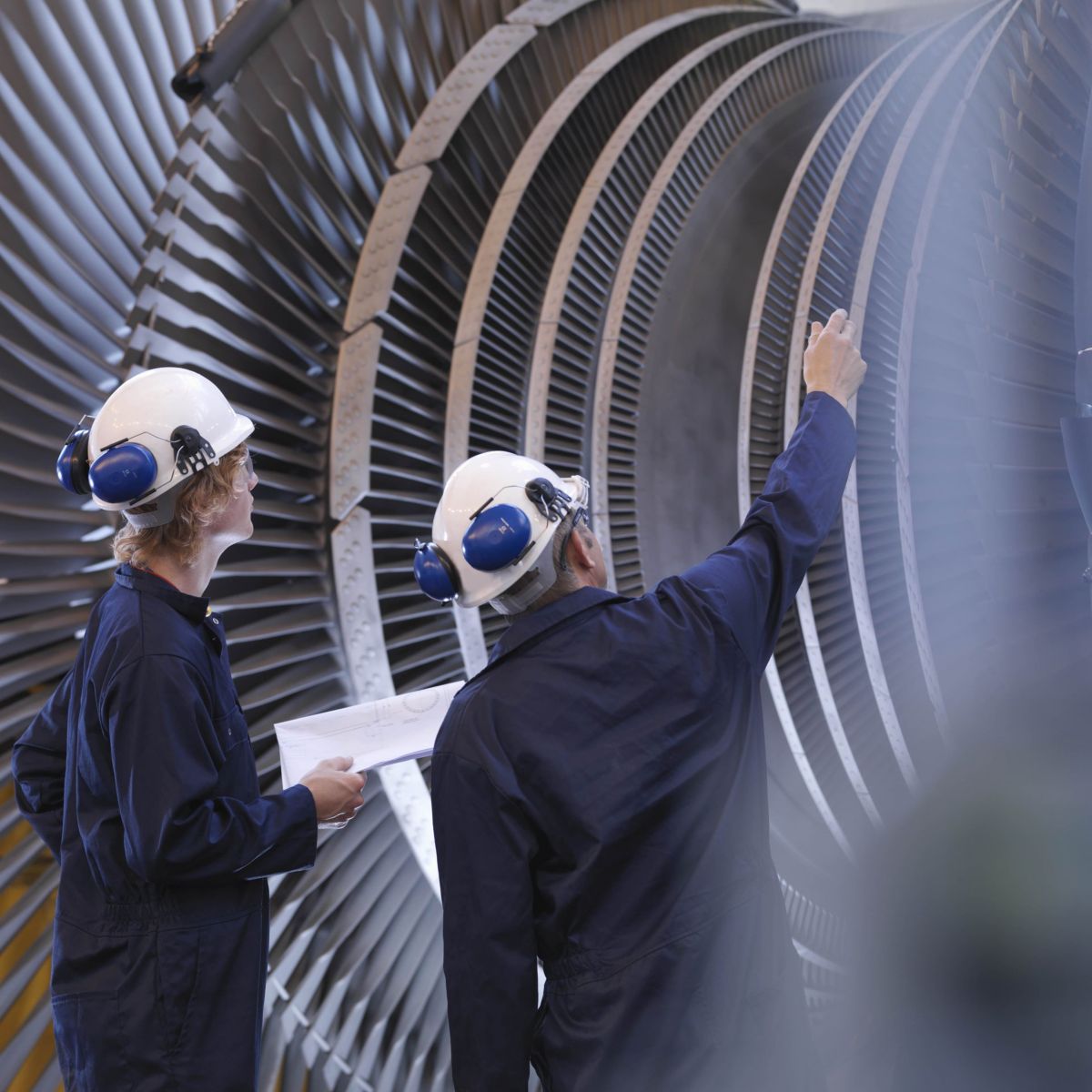 Two engineers in protective gear examine a massive industrial turbine rotor while reviewing technical schematics.