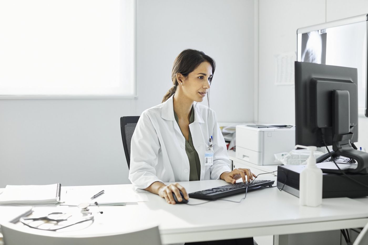 A professional female physician in a white coat works at her desk, using a computer in a clean, brightly lit hospital office environment.