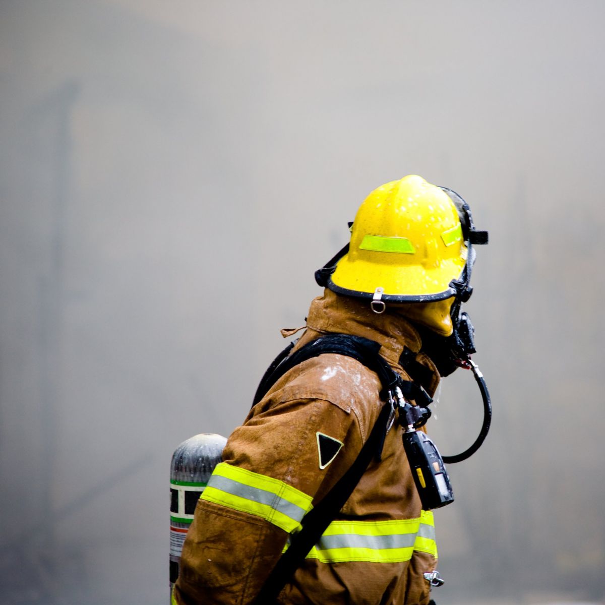 A firefighter in full protective gear and helmet navigates through dense, hazy smoke at the scene of an emergency.