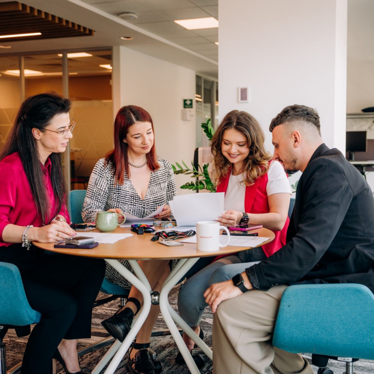 Four people seated around a table reviewing printed documents with laptops and personal items visible in an indoor office space.