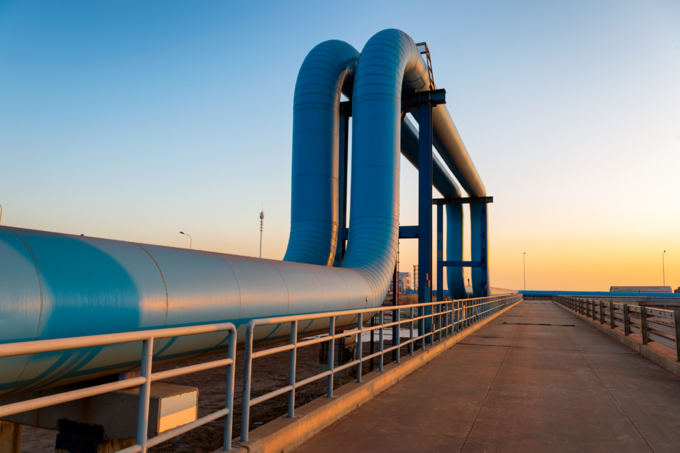 Massive blue pipes on a steel structure beside a concrete walkway under warm sunset light.