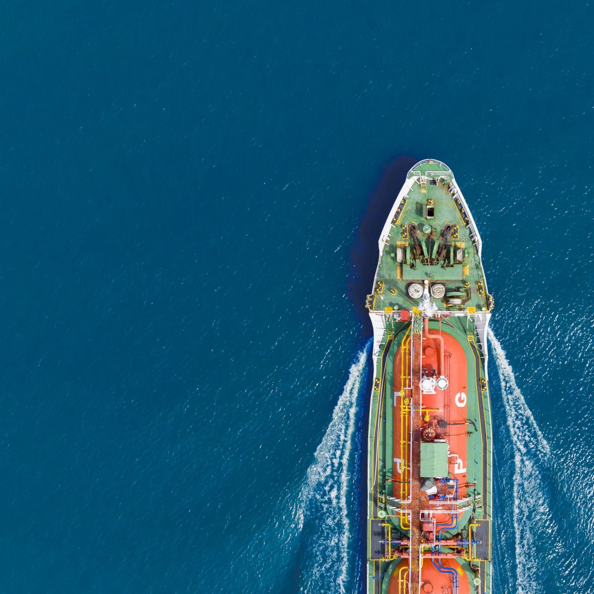 An aerial view captures a vibrant orange liquefied gas tanker moving through the deep blue ocean, leaving a white wake trailing behind.