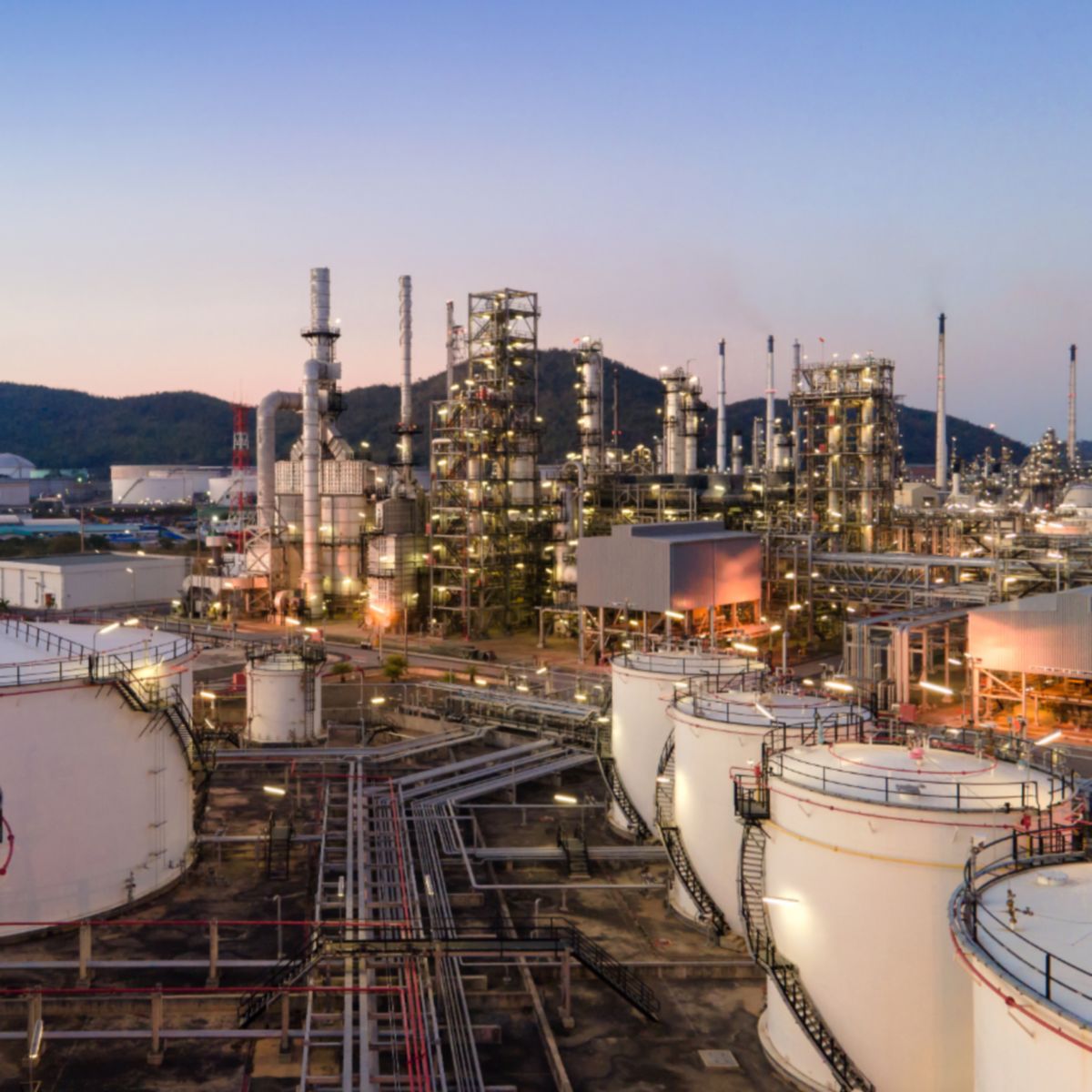 Industrial refinery complex with cylindrical tanks, pipes, and illuminated towers under an evening sky.