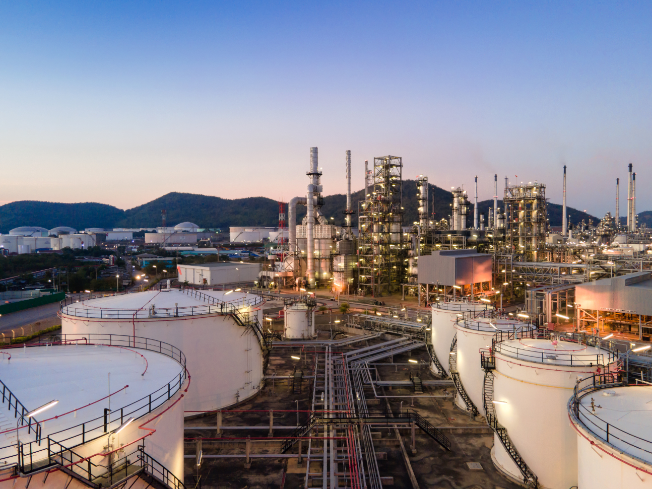 Industrial refinery complex with cylindrical tanks, pipes, and illuminated towers under an evening sky.