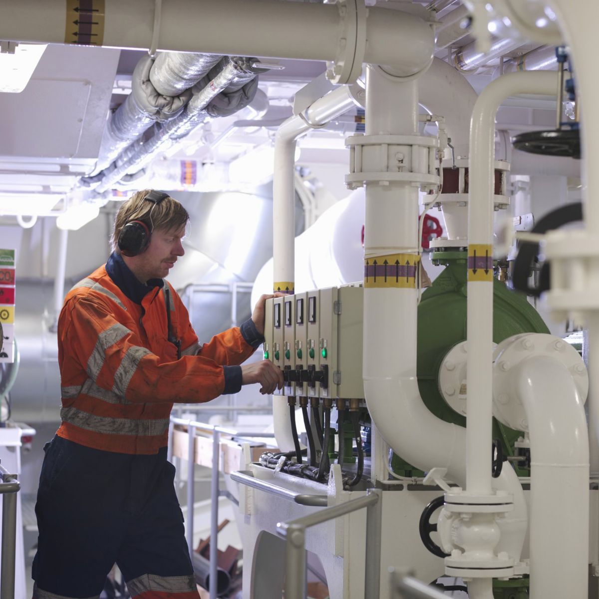 An industrial technician in high-visibility workwear operates machinery controls amidst a complex network of white pipes in a maritime engine room.