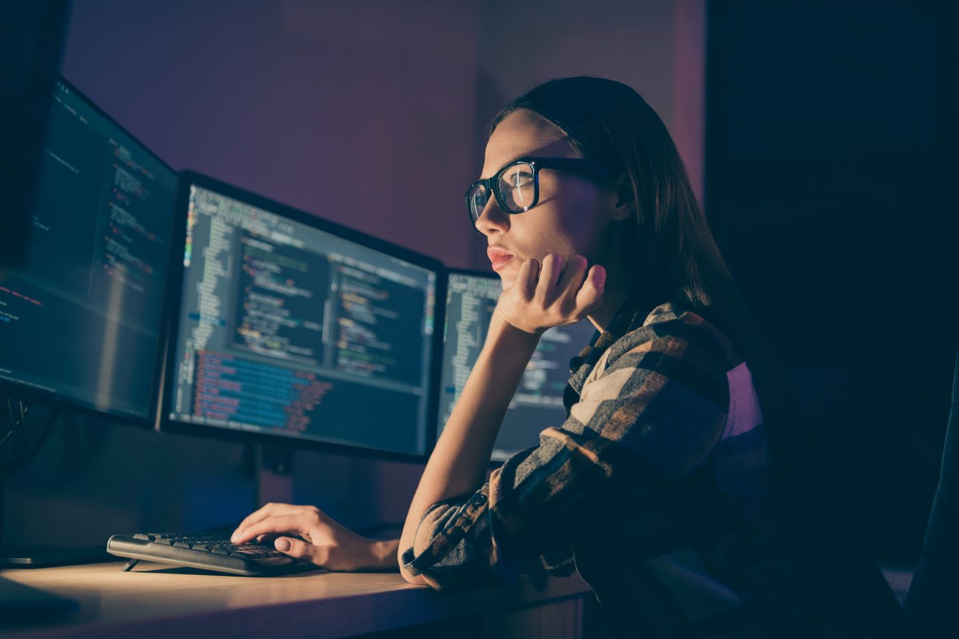 Person seated at a desk using a keyboard and viewing multiple monitors displaying code in a dimly lit indoor workspace.