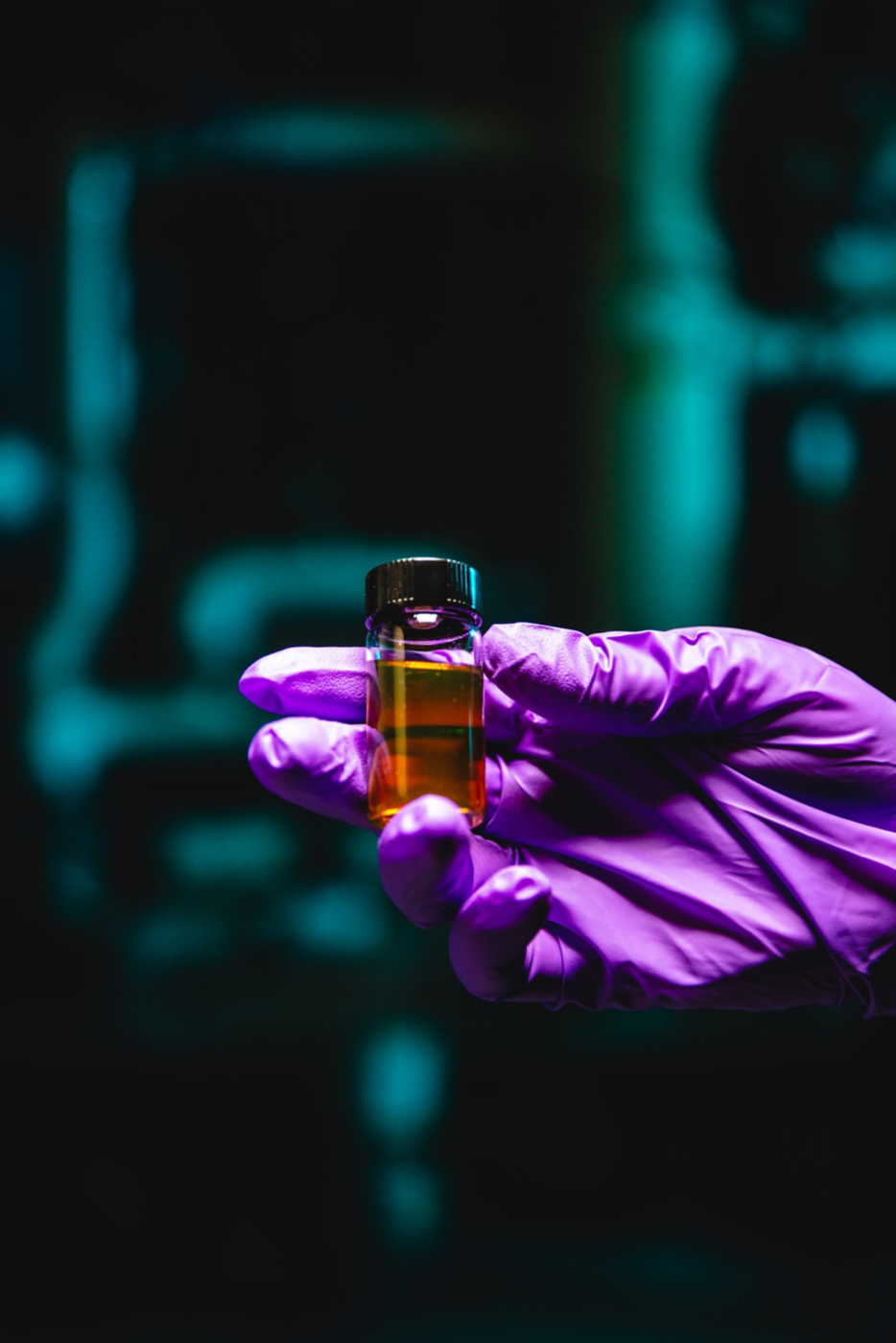 A scientist wearing a purple glove holds a small vial containing a yellow liquid against a dark, moody industrial background.