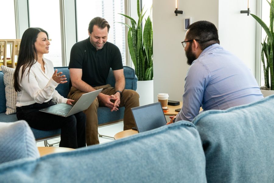 Three people sitting on chairs, one using a laptop, engaged in conversation in a room with plants, windows, and a small table.