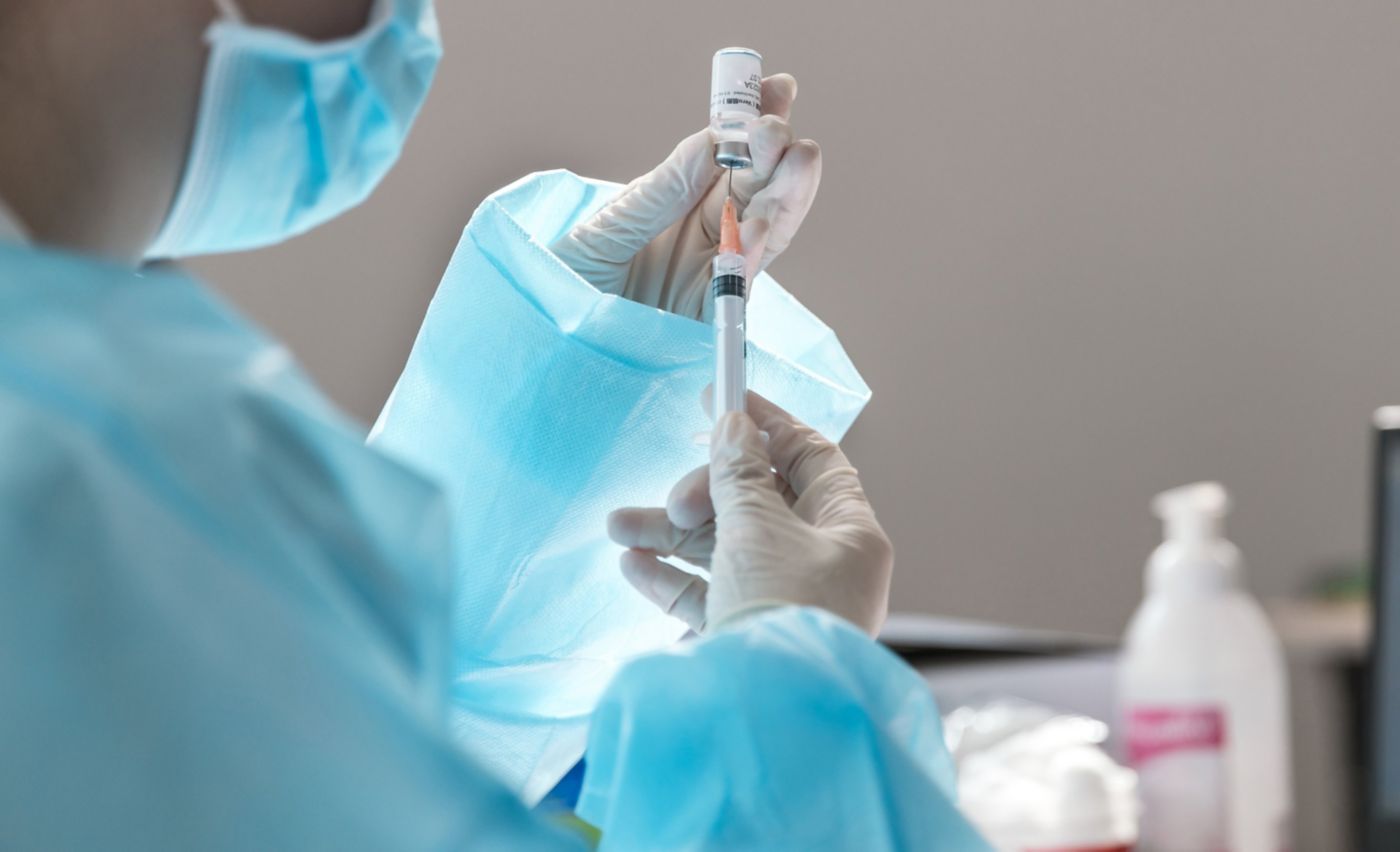 A medical professional in protective gear draws vaccine from a glass vial into a syringe for clinical administration.