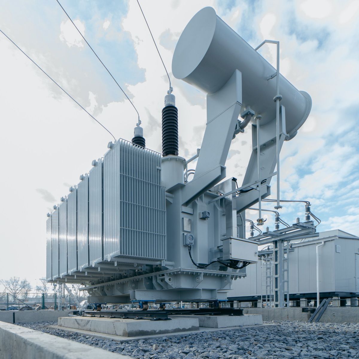 A large electrical transformer stands within an industrial substation under a bright, partly cloudy sky.