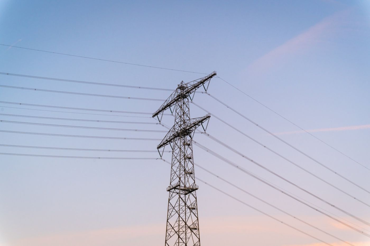 A tall metal transmission tower stands against a clear, soft-toned sky, showcasing complex electrical infrastructure in an industrial landscape.
