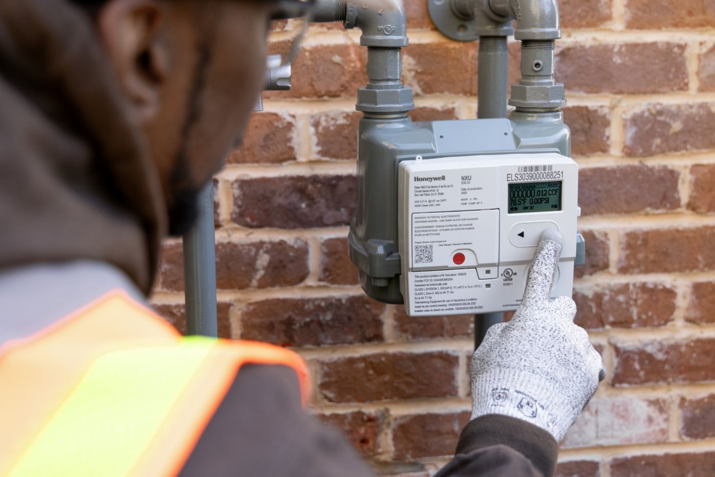 Person in Honeywell-branded safety clothing working with utility equipment installed among plants beside a brick building.