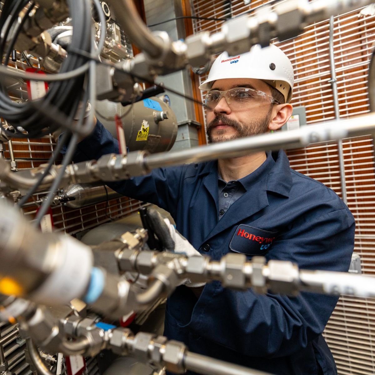 A focused technician in safety gear inspects complex industrial piping and control systems at a Honeywell facility.