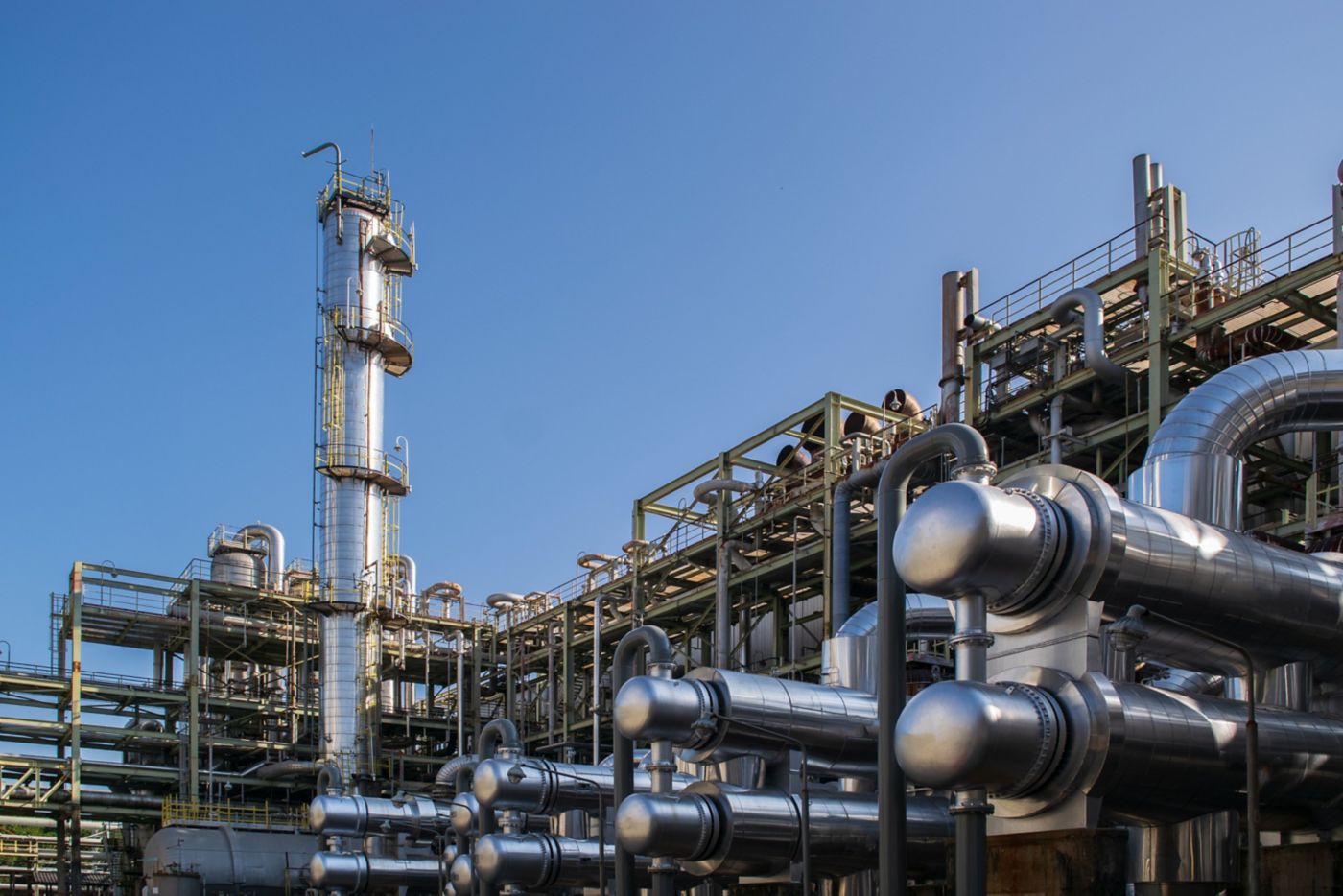 A complex array of insulated steel piping and distillation towers stands against a clear blue sky in an industrial chemical manufacturing facility.
