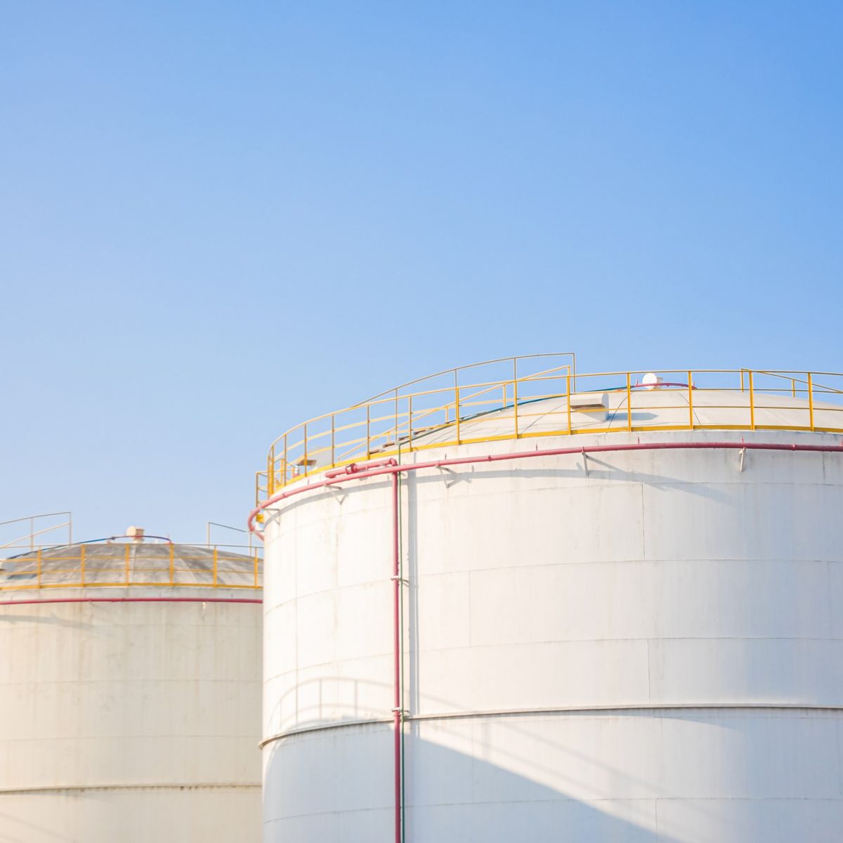 Large white industrial storage tanks against a clear blue sky, showing structural piping and safety railings for bulk chemical or fuel storage.