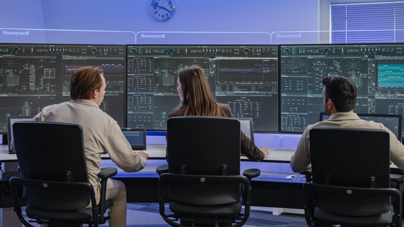 Three operators monitor complex data visualization screens in a high-tech corporate industrial control room environment.