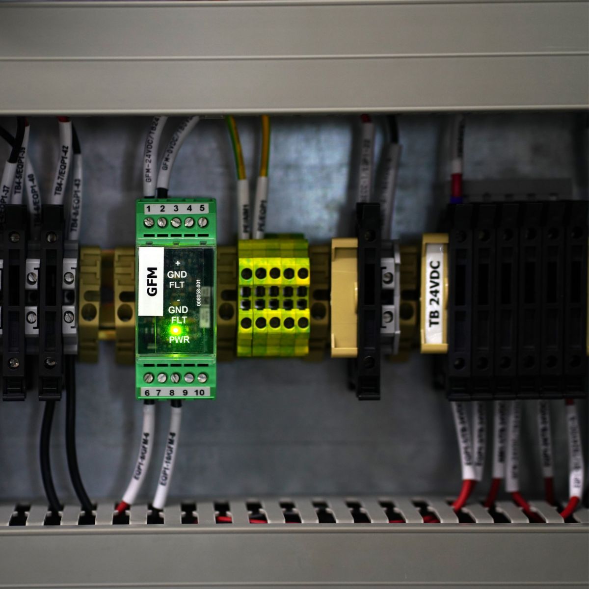 A close-up view of an active green LED GFM module mounted on a DIN rail within an industrial electrical control panel with organized cable labeling.